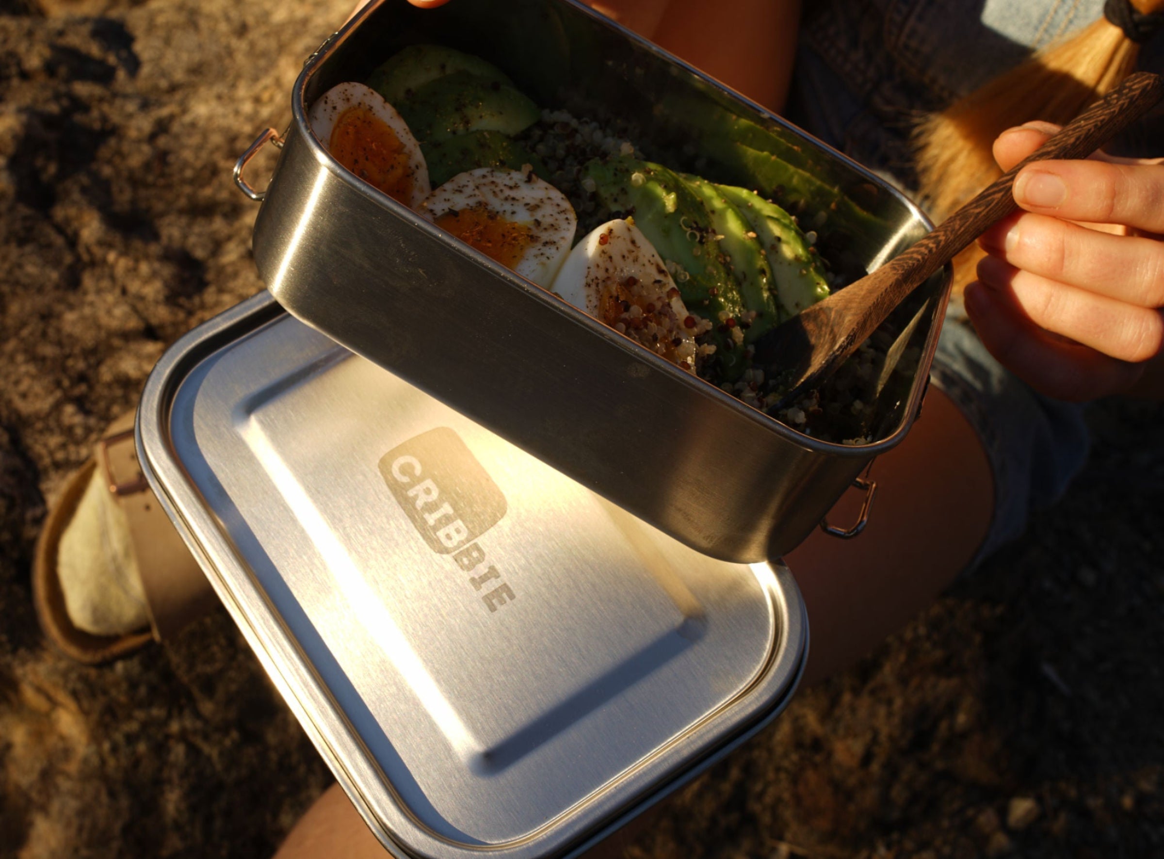 Stainless steel food container with food inside of it in nature with someone eating it with a spoon